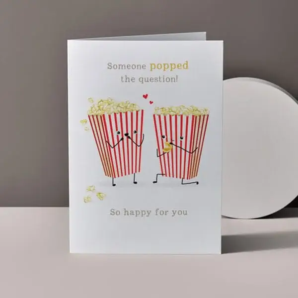 Engagement congratulations card featuring two illustrated popcorn boxes holding hands with popped the question message on a neutral background.